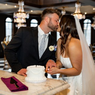 Bride and groom share a kiss while cutting a small white wedding cake on a marble bar inside an elegant reception space with crystal chandeliers, arched windows, and black-and-white checkered floors.